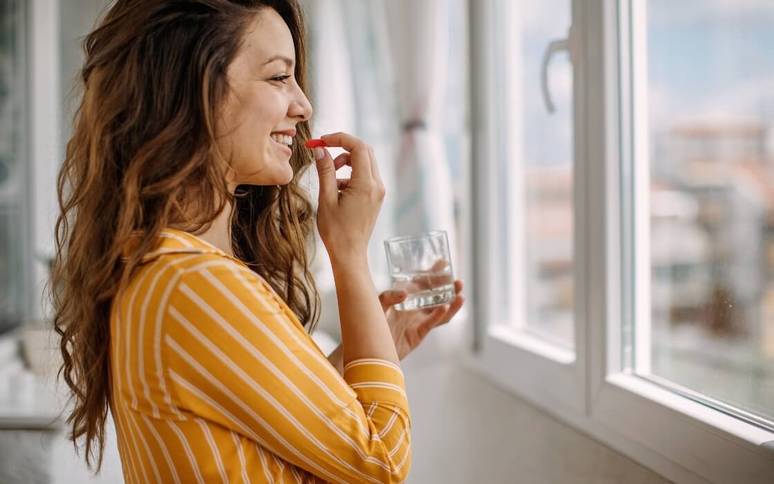 Frau nimmt am Fenster eine Kapsel mit einem Glas Wasser ein